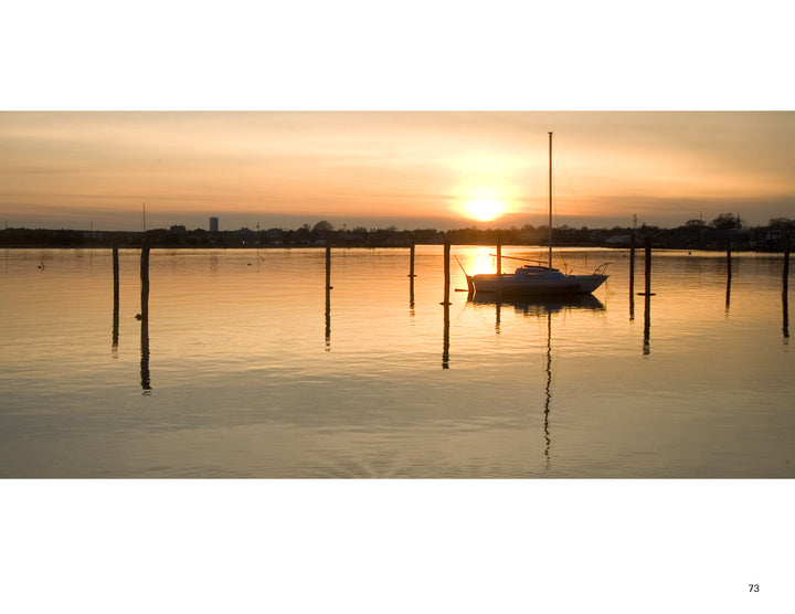 Martha’s Vineyard Wide by Schiffer Publishing, Ltd. captures a sailboat floating at sunset near Martha’s Vineyard, surrounded by wooden posts, with orange and yellow skies reflecting on calm waters and silhouettes of trees and buildings on the horizon.