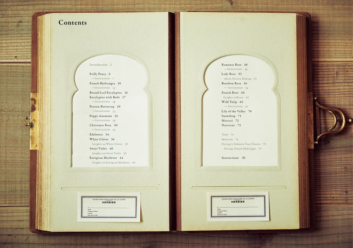 The Herbarium of Fabric Flowers by Schiffer Publishing, Ltd. lies open on a wooden table, showing its table of contents on textured cream pages with a gold clasp and two small cards at the bottom—ideal for herbarium fabric projects.