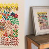 Split image: Left side displays a vibrant botanical embroidery on cream fabric. Right side features the same "Embroidery Garden" design by Schiffer Publishing, Ltd., framed and set on a wooden table against a plain white wall.