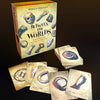 A box labeled Between the Worlds by Schiffer Publishing, Ltd. stands upright on a dark surface, surrounded by illustrated oracle cards depicting objects such as a ring, doll’s leg, horseshoe, sundial shell, and wishbone.