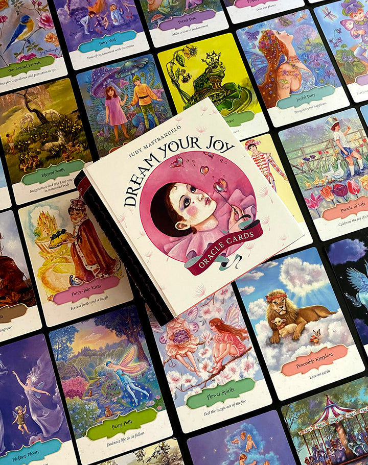 A box of Dream Your Joy Oracle Cards by Schiffer Publishing, Ltd. rests on vibrant affirmation cards, each showcasing whimsical, fantasy-inspired art and uplifting messages—ideal for dream meditation and positive inspiration.