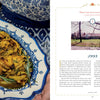 A person in a blue patterned apron holds a platter of roasted chicken with lemon wedges and herbs beside The Carlino Family Cookbook by Schiffer Publishing, Ltd., open to a page with family recipes and a house photo under the story titled 1995.