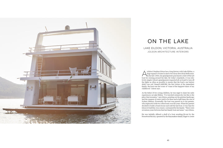 Second Home by Schiffer Publishing, Ltd. is a modern, architect-designed houseboat with outdoor seating on a calm lake. Large windows and a white haven interior offer sanctuary and rejuvenation in a serene, luxurious setting surrounded by hills.