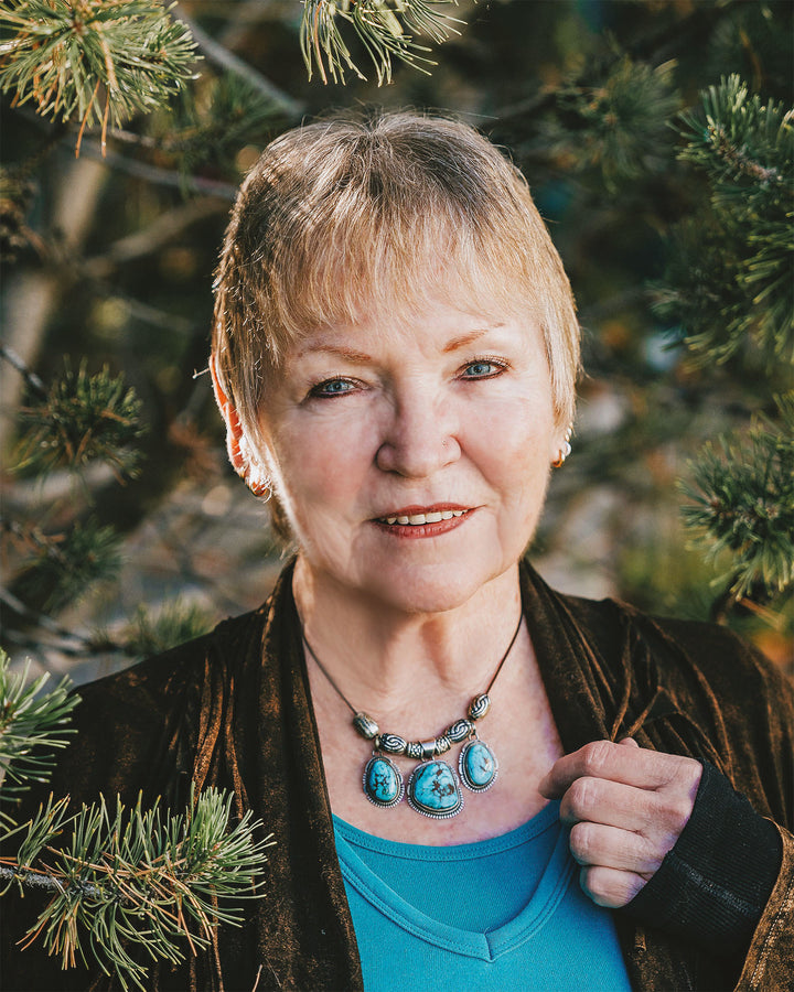 An older woman with short blonde hair stands outdoors among pine branches, smiling gently at the camera while holding the Steele Wizard Tarot by Schiffer Publishing, Ltd. She's dressed in a turquoise top, brown jacket, and statement turquoise necklace.