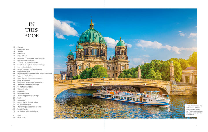 A scenic city view features a large domed cathedral beyond a stone bridge over a river, where a white tour boat from "River Cruises" by Schiffer Publishing, Ltd. glides beneath, framed by trees and a blue, cloud-streaked sky.