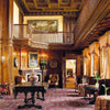In "At Home with Royalty" by Schiffer Publishing, Ltd., experience a grand castle room with wood paneling, gold-curtained windows, antique furniture, and a suited man near the fireplace below a balcony adorned with artwork.