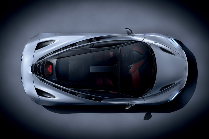 Overhead view of McLaren by Schiffer Publishing, Ltd.—a sleek silver car with a glass roof and visible red interior—photographed on a dark, spotlighted background.
