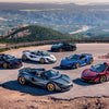 Six McLaren supercars by Schiffer Publishing, Ltd. are parked on a winding mountain road, overlooking a scenic valley with forests, lakes, and distant hills under a partly cloudy sky.