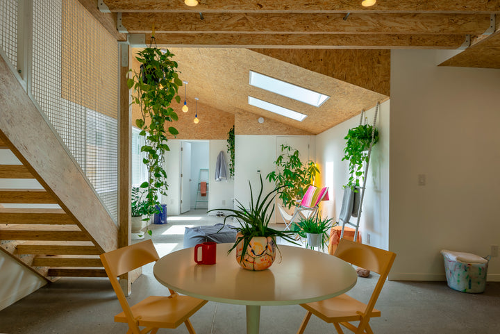 A stylish small room from "The Well-Designed Accessory Dwelling Unit" by Schiffer Publishing, Ltd. features a round table, yellow chairs, hanging plants, large windows, a wooden staircase, and sunlight brightening cozy green seating.