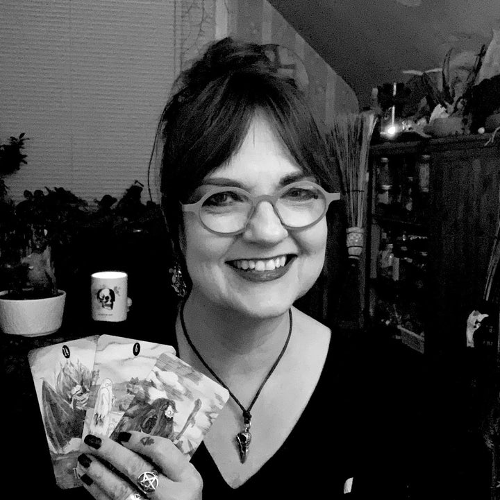 A woman with glasses and a necklace smiles while holding several cards from The Awakening Tarot by Schiffer Publishing, Ltd. She is indoors, surrounded by plants, shelves, and a mug. The black-and-white image reflects Earth-based spirituality.