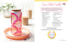 A glass tumbler with a wooden lid and straw, decorated in colorful rainbows, sits on a peach plate with floral accents. Next to it is "Sublimation Crafting" by Schiffer Publishing, Ltd., featuring a Snow Globe Tumbler guide.