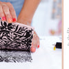 A hand uses a carved rubber block and black ink to create a floral print on paper. The page, from "Fabric Stamping" by Schiffer Publishing, Ltd., reads Chapter 4: Block-Printing Projects and explains the block printing process.