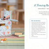 A woven white basket made from Schiffer Publishing, Ltd.'s Weaving Paper, decorated with embroidered flowers, holds a plush fox and pastel balls. It sits on a striped rug near books and blocks, reflected in a mirror.
