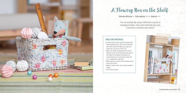 A woven white basket made from Schiffer Publishing, Ltd.'s Weaving Paper, decorated with embroidered flowers, holds a plush fox and pastel balls. It sits on a striped rug near books and blocks, reflected in a mirror.
