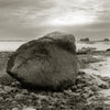 A large, weathered boulder sits on a rocky shore beneath cloudy skies in "The Maine Coast" by Schiffer Publishing, Ltd., evoking tranquil moods with distant trees and calm water in the background.