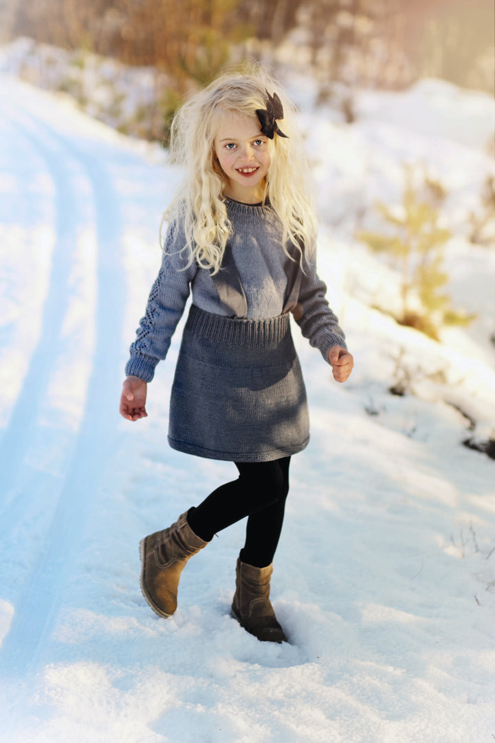 A young child wearing a Nordic-style grey dress from "Winter Knitting for Little Sweethearts" by Schiffer Publishing, Ltd. walks on a snowy path, dressed with black leggings, brown boots, and a black hair bow amid winter trees.