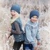 Two children in blue Nordic-inspired knit hats and sweaters, from "Winter Knitting for Little Sweethearts" by Schiffer Publishing, Ltd., stand smiling in a frosty field with wintry grasses, looking back at the camera.