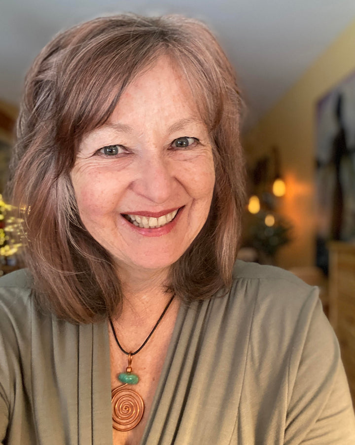 A smiling woman with shoulder-length light brown hair wears a green top and a spiral pendant, standing indoors with warm lighting that reflects the earth-centered spirituality of the Gaian Tarot by Schiffer Publishing, Ltd.