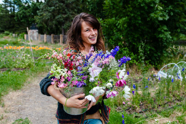 A woman stands in a vibrant garden with a bouquet, embodying the DIY spirit featured in "Sow, Grow, and Harvest" by Schiffer Publishing, Ltd. Lush trees and blooming plants surround her on this sunny day—perfect inspiration for flower enthusiasts.