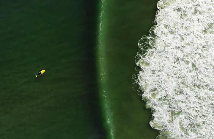 Out East from Above by Schiffer Publishing, Ltd. features aerial photography of a lone surfer on a yellow board paddling toward a large green wave off South Fork Long Island, with white foamy surf breaking to the right.