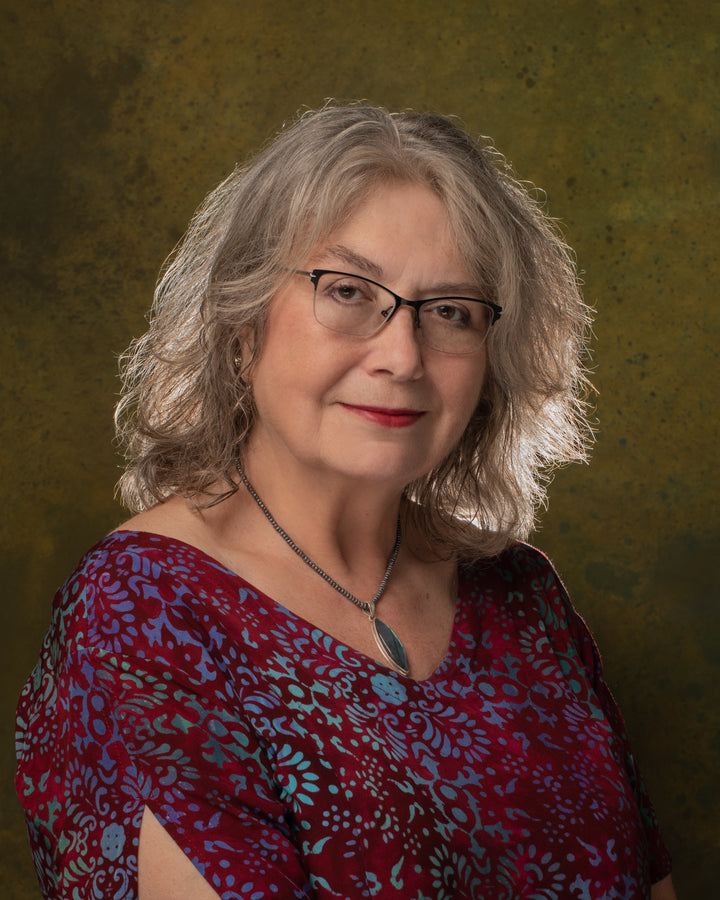An older woman with wavy gray hair, glasses, a red patterned top, and a silver necklace with blue pendant poses like a Boadicea's Tarot of Earthly Delights card by Schiffer Publishing, Ltd. against a mottled green and brown backdrop.