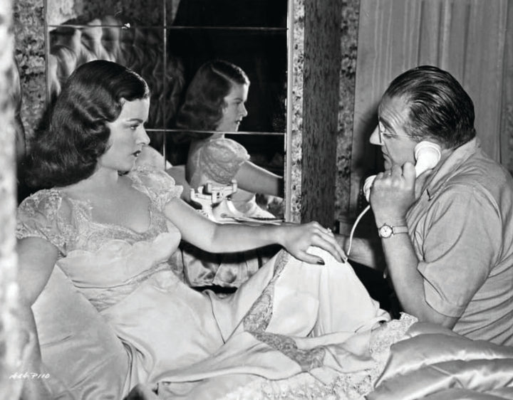 A woman in a nightgown sits on a bed as a man talks on the phone—a scene echoing The Art of Classic Crime and Mystery Movies by Schiffer Publishing, Ltd., with a mirror, drapes, and wallpaper creating a cinematic backdrop.