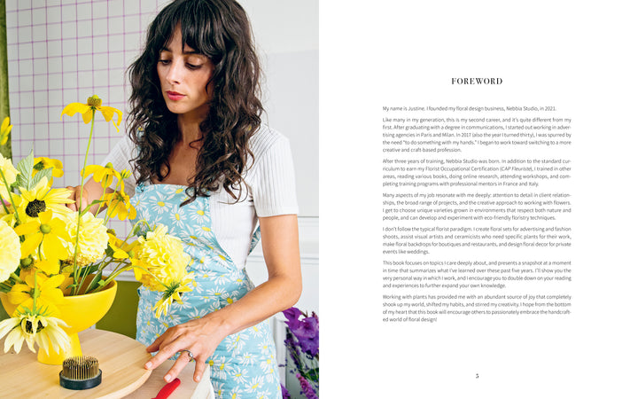 A woman with long dark hair, wearing a light blue floral dress over a white top, arranges yellow flowers on a wooden table beside "Starting Your Floral Design Studio" by Schiffer Publishing, Ltd., opened to its foreword page.