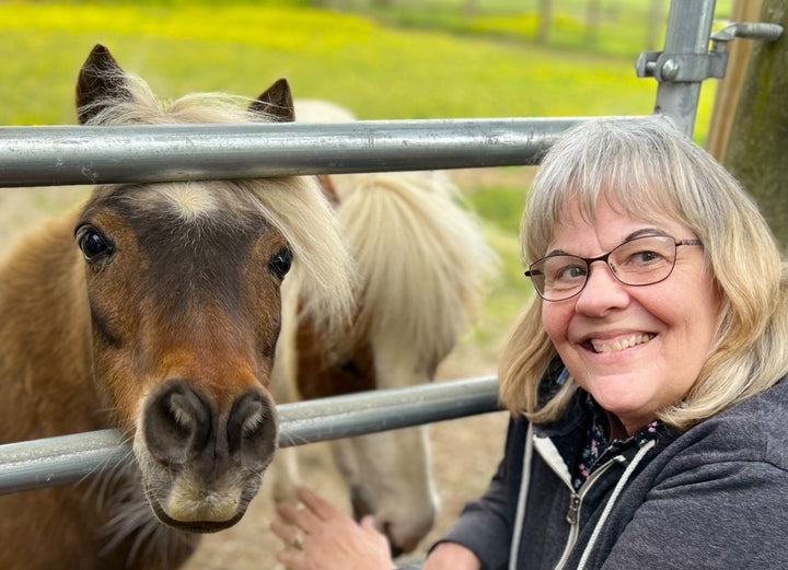 A smiling woman with glasses and gray hair poses next to a small brown and white Chincoteague pony behind a metal fence outdoors, holding a copy of “Out of the Sea” by Schiffer Publishing, Ltd.