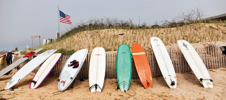 Surfboards of various colors and sizes stand in sand before grassy dunes and an American flag—a classic Montauk scene featured in Schiffer Publishing, Ltd.’s "Montauk 11954, 2nd Edition.