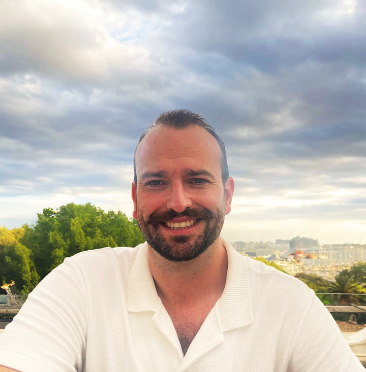 A man with short brown hair and a beard, wearing a white polo, smiles outdoors amid trees and a cloudy harbor, capturing the self-discovery theme of Lieselle's Eternal Tarot by Schiffer Publishing, Ltd.