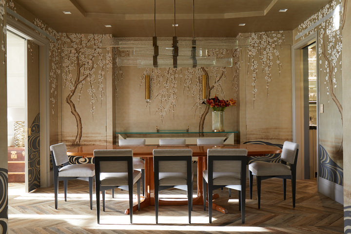 A dining room inspired by Penny Drue Baird, as featured in "PowerHouse" by Schiffer Publishing, Ltd., with a wooden table, eight upholstered chairs, a floral mural, glass chandelier, chevron floor, and a side table with a vase. Light streams in from the left.