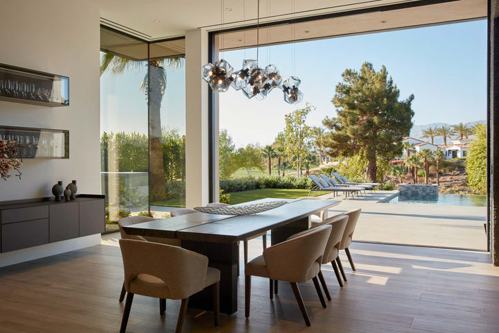 The PowerHouse dining room by Schiffer Publishing, Ltd. showcases beige chairs and a dark wood table, large glass doors to a sunny patio, pool, garden with mountain views, and geometric pendant lights.