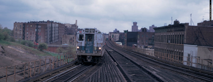 A graffiti-covered subway car travels on a weathered elevated track past city apartments, evoking NYC history as seen in "New York Subways and Stations, 1970–1990" from Schiffer Publishing, Ltd.