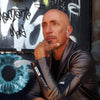 A bald man in a leather jacket sits thoughtfully before a graffiti wall and painted blue eye, evoking the vivid, surreal style of the Starman Tarot Remastered by Schiffer Publishing, Ltd., inspired by David Bowie.