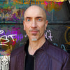 A bald man with blue eyes, a trimmed goatee, and earrings wears a dark brown textured jacket, standing before a vibrant graffiti wall inspired by Schiffer Publishing, Ltd.'s Starman Tarot Remastered.