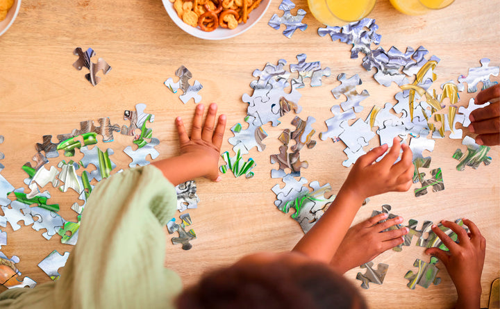 Two children assemble The Tiniest Giant 4-in-1 Puzzle Set by Schiffer Publishing, Ltd. on a wooden table covered with puzzle pieces, with snacks and juice on the side.
