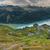 A panoramic view of winding mountain roads and green hills above a blue lake evokes the dramatic scenery featured in "Alpine Railways" by Schiffer Publishing, Ltd., reminiscent of classic European Alps train journeys.