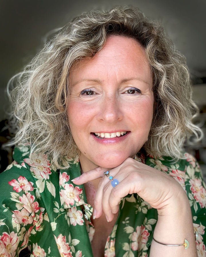 A smiling woman with curly blonde hair and a green floral shirt rests her chin on her hand, wearing a blue stone ring. She sits in soft natural light, ready to share "Yummy Watercolor" by Schiffer Publishing, Ltd. for beginners.