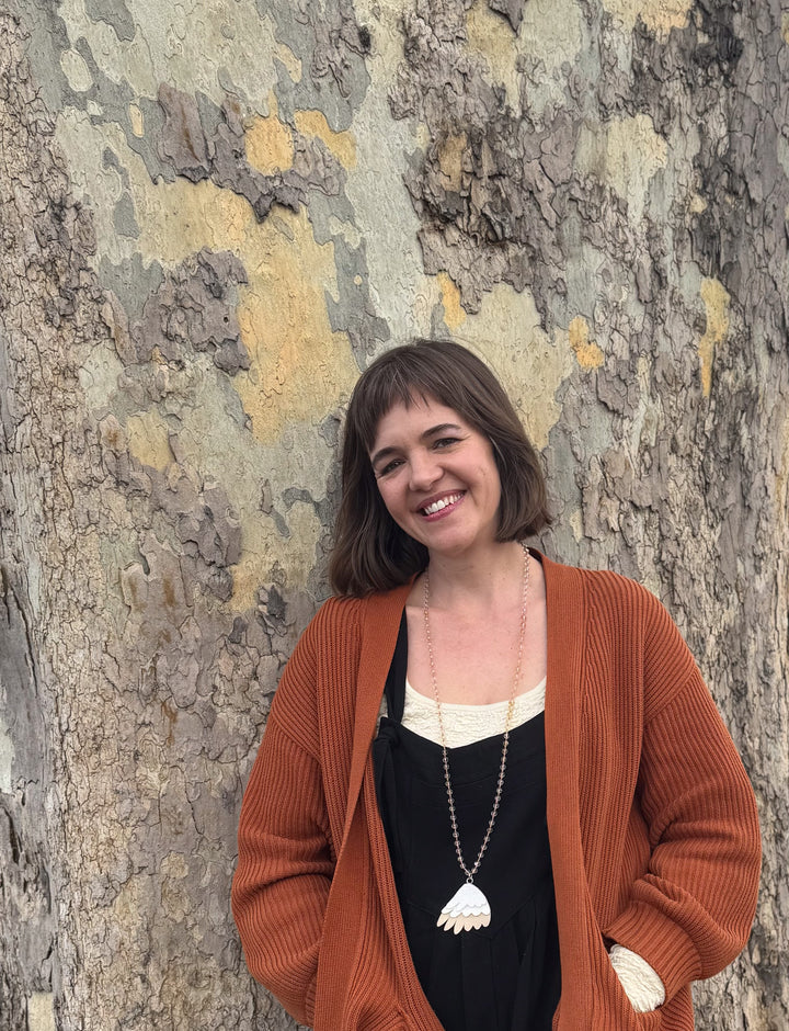 A woman with short brown hair and an orange cardigan smiles by a tree trunk—capturing a scene perfect for "The World in Me" by Schiffer Publishing, Ltd.