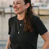 A woman with long brown hair, wearing a black t-shirt and jewelry, smiles by the marina at sunset, holding "Mermaid Candy" by Schiffer Publishing, Ltd., as if treasuring sea glass from a mermaid story.