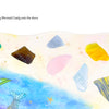 Mermaid tails rise from blue waves near a sandy shore sparkling with sea glass and golden stars. Text: At dawn, Heart scattered Mermaid Candy by Schiffer Publishing, Ltd. along the shore in this magical mermaid picture book.