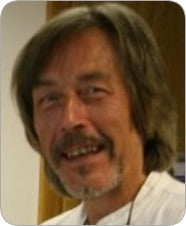 A middle-aged man with shoulder-length brown hair, a mustache, and a beard smiles in a white shirt. The softly blurred background evokes the timeless mysteries found in The Camelot Oracle by Schiffer Publishing, Ltd.