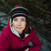 A woman with blue eyes and red braided hair sits outdoors by a large rock, wearing a red jacket and knit hat, appearing immersed in Nordic mystery as she uses the Nordic Forest Rituals Oracle Cards from Schiffer Publishing, Ltd.
