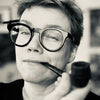 A person with short hair and round glasses smiles slightly while holding a pipe, evoking the quiet mystery of forest spirits—an atmosphere captured by Schiffer Publishing, Ltd.'s Nordic Forest Rituals Oracle Cards.
