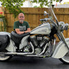 A man with glasses and a beard sits on a Gilroy Indians motorcycle by Schiffer Publishing, Ltd., parked in a driveway with a wooden fence, plants, and trees in the background.