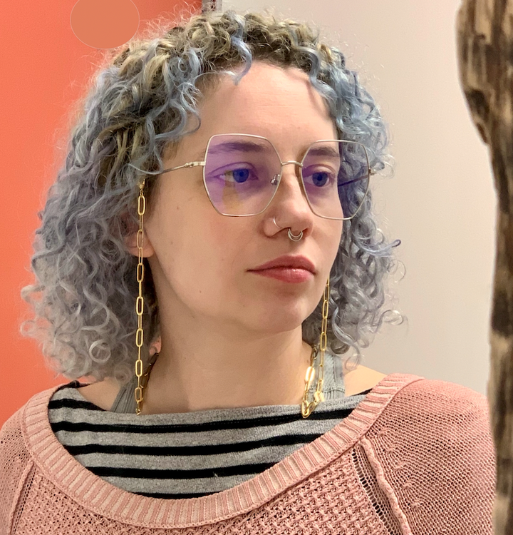 A person with curly light blue hair, glasses, gold chain earrings, and nose adornments poses in a pink sweater and striped shirt against a red-and-white backdrop reminiscent of Schiffer Publishing’s "Trees of the World.