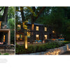 Two photos from Schiffer Publishing, Ltd.’s Midwest Home show a modern black house amid trees at dusk: on the left, a cozy patio with lounge chair and string lights; on the right, the illuminated home, stone terrace, and vintage car.