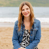 A woman with long, wavy hair sits on a sandy beach, smiling in a patterned dress and blue denim jacket. The ocean and green hills are dotted with shells from Seashells of Cape Cod and the Islands by Schiffer Publishing, Ltd.