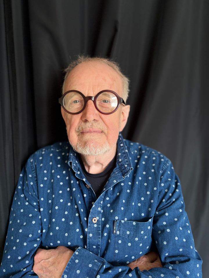 An older man with a gray beard and round glasses stands with arms crossed in a blue polka-dot shirt before a black curtain, embodying the spirit of "The Story of the Blues in 50 Songs" by Schiffer Publishing, Ltd.