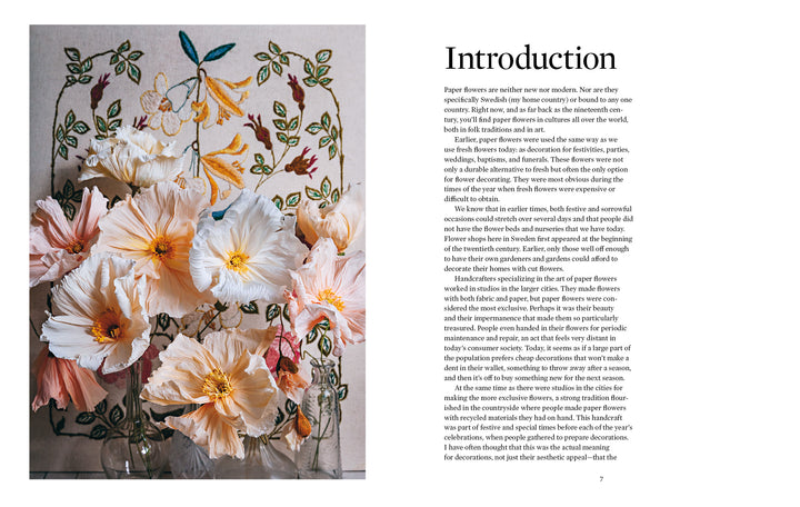 The book "Paper Flower Magic" by Schiffer Publishing, Ltd. rests beside pastel crepe-paper blooms in a glass vase, placed before an embroidered floral wall hanging and a book page titled Introduction about paper flowers.